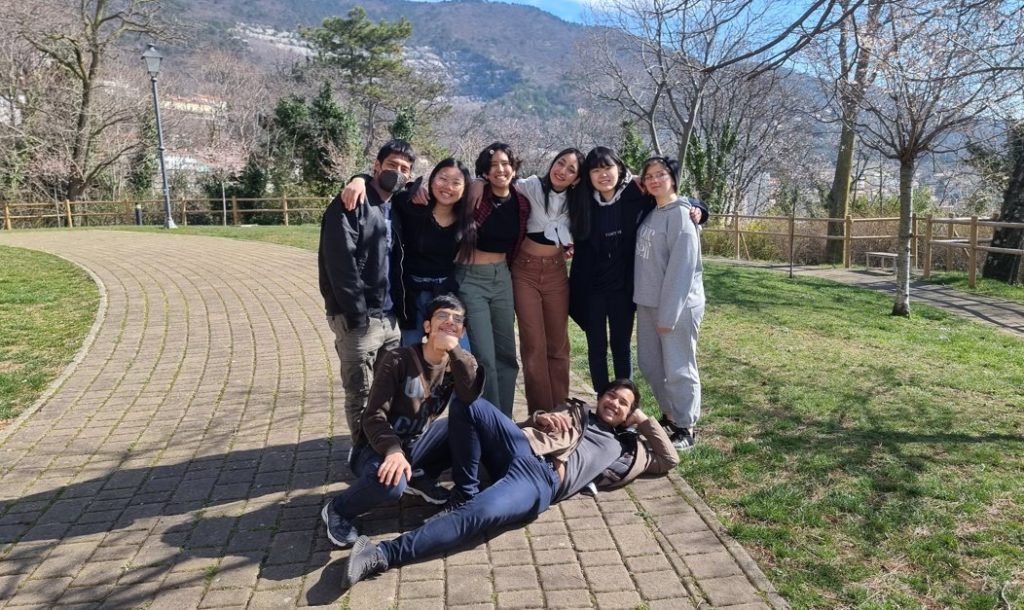 A group of nine people is gathered on a sunlit path in a park. They are smiling and embracing each other, with some sitting and others standing. Leafless trees and hills are visible in the background. It appears to be a bright, clear day.