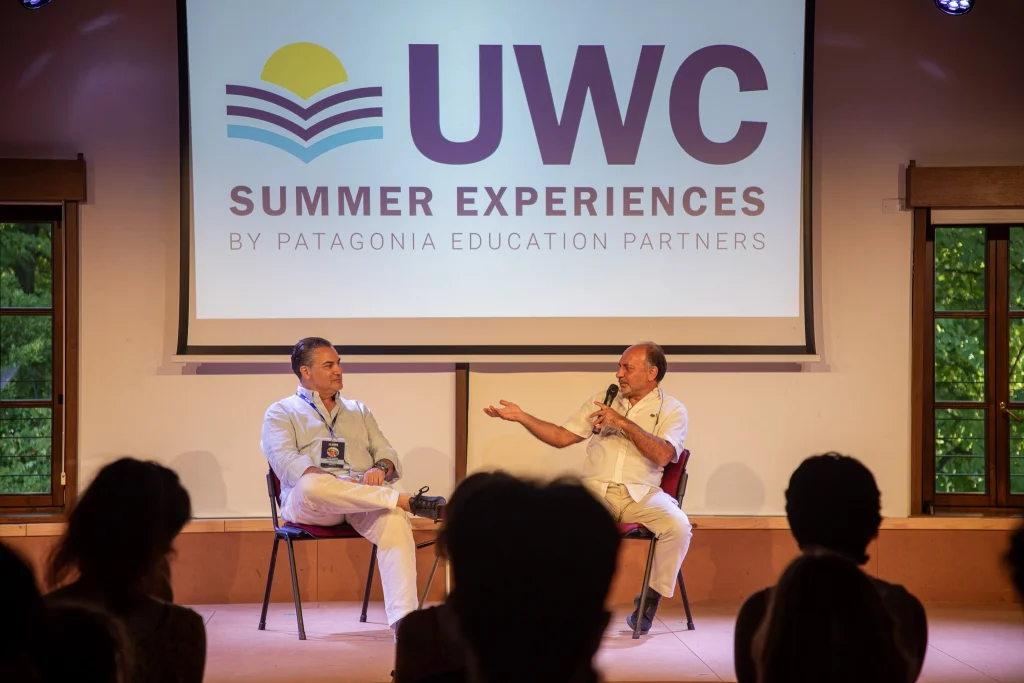 Due uomini siedono sul palco e discutono davanti a uno schermo con il logo "UWC Summer Experiences by Patagonia Education Partners", mentre il pubblico ascolta.