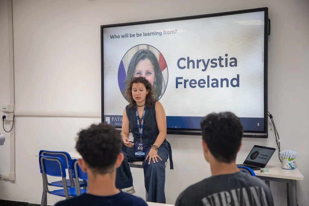 Una donna siede su un banco di fronte a un'aula e parla agli studenti. Dietro di lei, un grande schermo mostra una foto e il nome "Chrystia Freeland", insieme al testo "Da chi impareremo?".