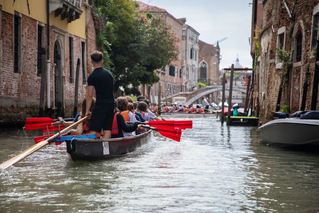 Persone remano con remi rossi su barche di legno lungo uno stretto canale fiancheggiato da vecchi edifici in mattoni a Venezia, Italia. Altre barche e un ponte pedonale in pietra sono visibili sullo sfondo sotto un cielo parzialmente nuvoloso.