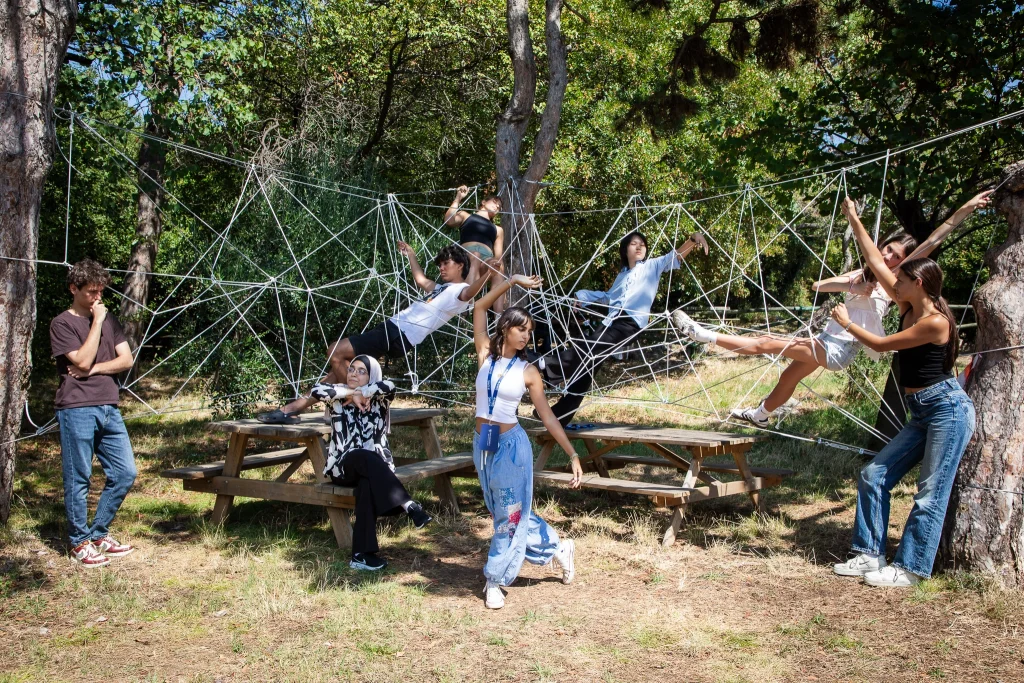 Un gruppo di giovani interagisce con una grande rete di corde bianche tesa tra gli alberi in un'area boschiva, alcuni in piedi, altri seduti su una panchina, altri ancora arrampicati o appesi alle corde.