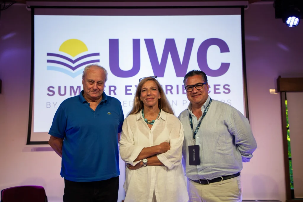 Tre persone sono in piedi, sorridenti, davanti a uno schermo che mostra il logo di UWC Summer Experiences. I due uomini portano gli occhiali, uno indossa una polo blu e l'altro una camicia chiara. La donna al centro indossa una camicetta bianca.