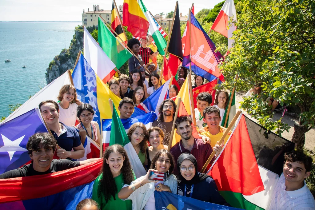 Un gruppo eterogeneo di giovani sorridenti tiene in mano e sventola diverse bandiere nazionali su una passerella soleggiata e panoramica che si affaccia sul mare e sulle scogliere, con alberi e un edificio visibili sullo sfondo.