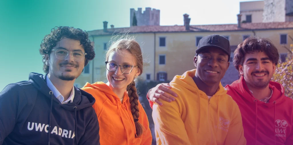 Four smiling young adults sit outdoors, wearing colorful hoodies with "UWC ADRIATIC" logos. Two men and a woman pose with arms around each other, with historic buildings and trees in the background under a clear sky.