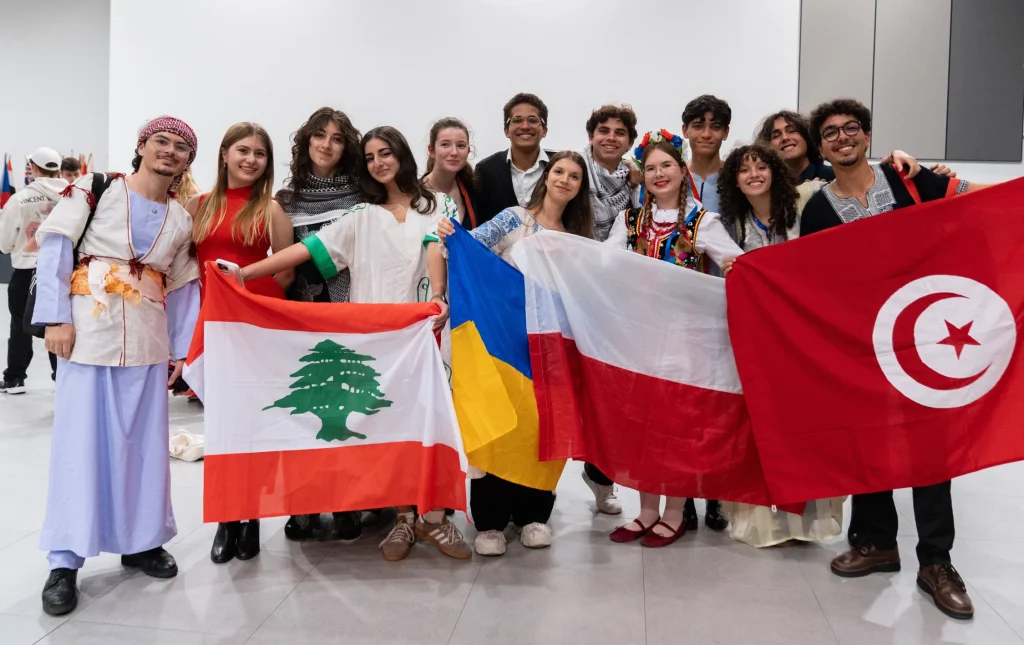 Un gruppo di giovani sorridenti, vestiti con abiti tradizionali diversi, si trova al chiuso con in mano le bandiere di Libano, Ucraina, Polonia e Tunisia, per celebrare la diversità culturale.