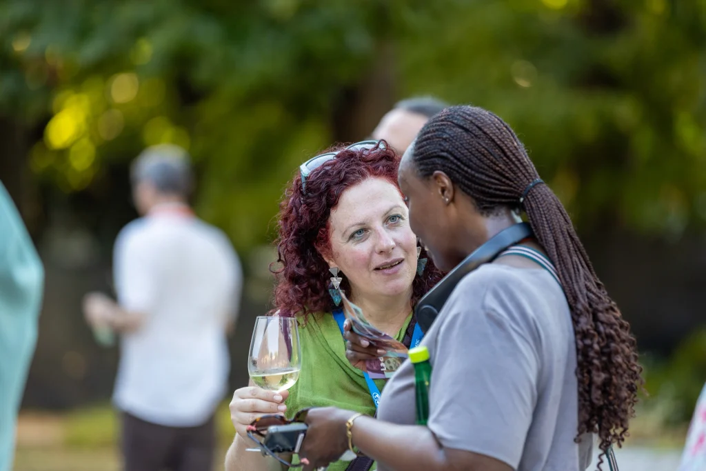 Due donne conversano all'aperto durante un evento sociale; una delle due tiene in mano un bicchiere di vino bianco. Entrambe sembrano intente a parlare. Sullo sfondo si intravedono altre persone sfocate, con gli alberi che fanno da cornice verde.