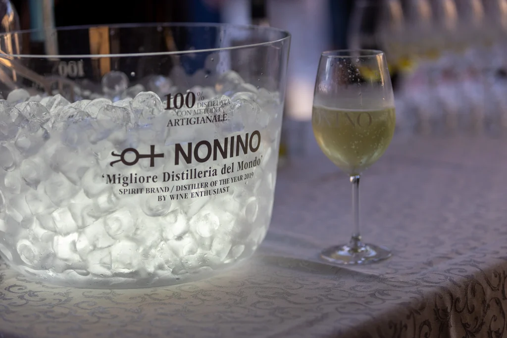 Su un tavolo, accanto a un bicchiere di vino bianco, c'è una secchiella trasparente per il ghiaccio con la scritta "Nonino", piena di ghiaccio. La secchiella reca una scritta in italiano ed è illuminata dalla luce del sole.