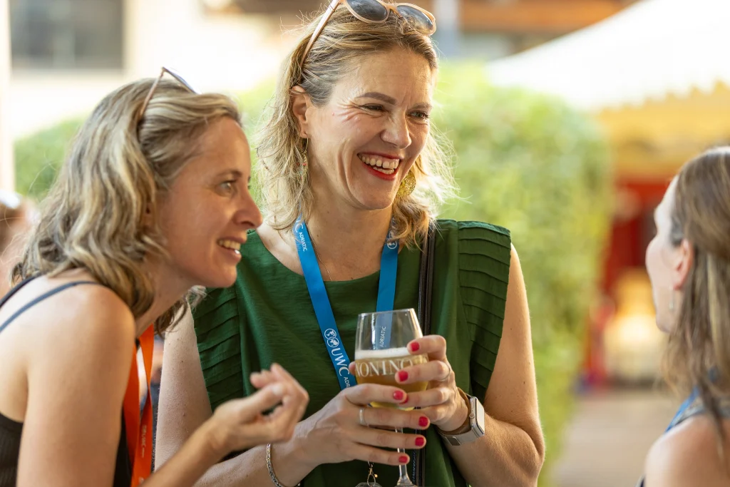 Tre donne stanno chiacchierando e ridendo all'aperto. Una di loro ha in mano un drink e porta gli occhiali da sole sulla testa. Sembrano tutte godersi una conversazione amichevole in una giornata di sole.