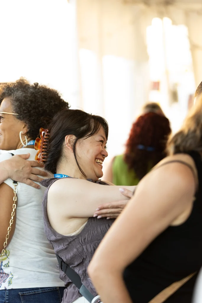 Due donne si abbracciano e sorridono in un ambiente interno ben illuminato. Sullo sfondo si intravedono altre persone, leggermente sfocate. L'atmosfera sembra calorosa e amichevole.