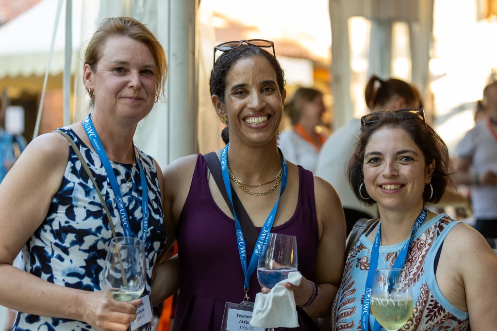 Tre donne con badge identificativi e cordoncini blu sorridono alla fotocamera, ciascuna con un bicchiere di vino in mano, durante un evento all’aperto con persone sfocate e tende sullo sfondo.