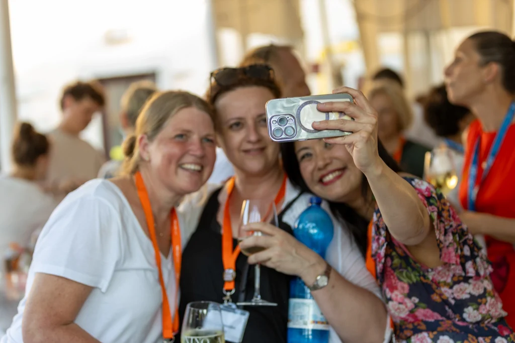 Tre donne con dei cordoncini arancioni sorridono e posano per un selfie durante un evento sociale, circondate da altre persone sullo sfondo. Una delle donne tiene in mano un telefono e tutte sembrano allegre e rilassate.
