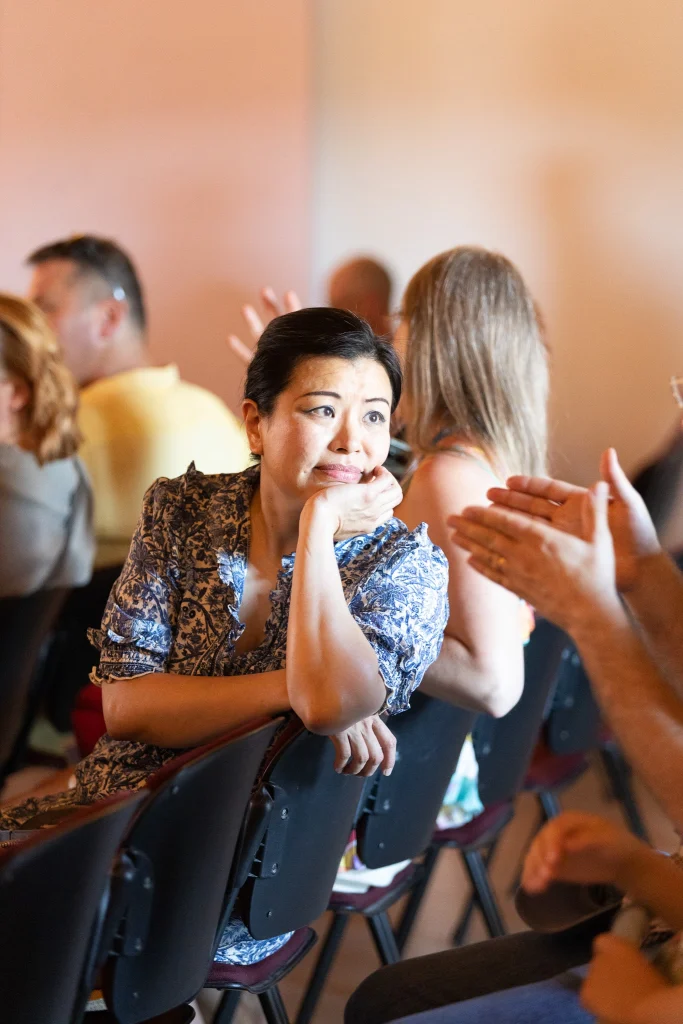 Una donna è seduta in mezzo a un gruppo di persone, con il mento appoggiato sulla mano e l'aria pensierosa, mentre gli altri intorno a lei conversano e applaudono.