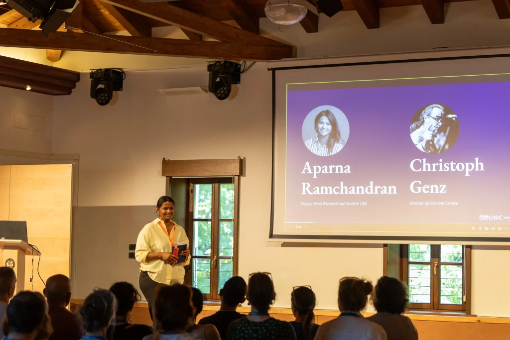 Una donna è in piedi vicino a un podio e sta parlando al pubblico. Dietro di lei, uno schermo mostra foto e nomi: Aparna Ramchandran e Christoph Genz. La sala ha travi a vista e è illuminata dalla luce naturale che entra dalle finestre.