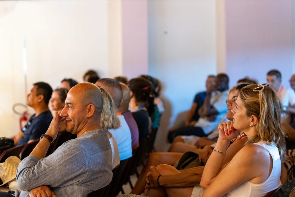 Un gruppo di persone sedute al chiuso, che osservano con attenzione qualcosa fuori campo. Alcune sorridono e sembrano coinvolte, mentre nell'ambiente si respira un'atmosfera rilassata.