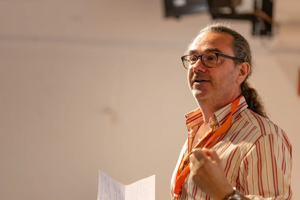 Un uomo con gli occhiali e una maglietta a righe parla tenendo in mano dei fogli e indossando un cordino arancione. Lo sfondo è sfocato, il che fa pensare a un evento o a una presentazione al chiuso.