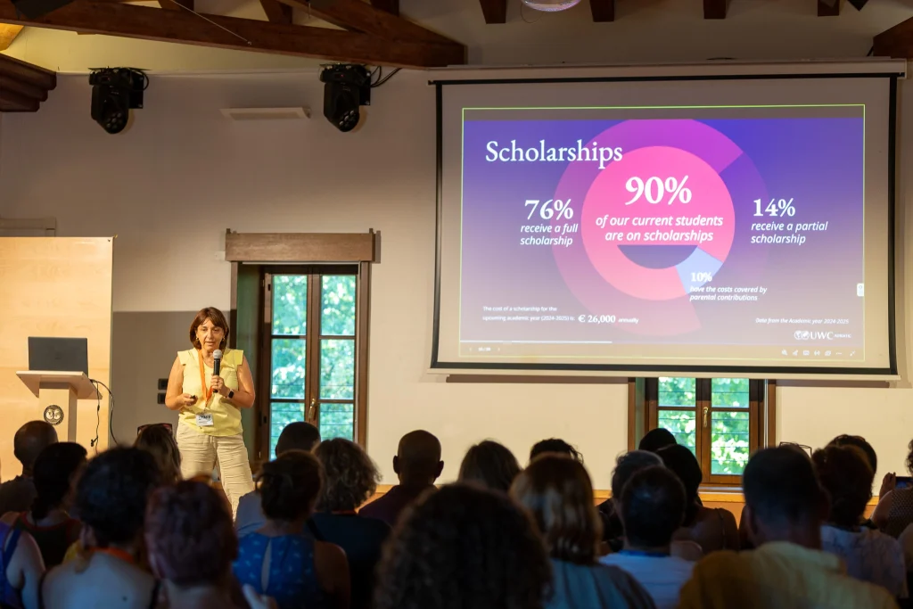 Una donna tiene una presentazione davanti a un pubblico, mostrando una diapositiva con dati statistici sulle borse di studio, tra cui: «Il 90% degli studenti attuali beneficia di una borsa di studio». La sala ha travi a vista e due grandi finestre.