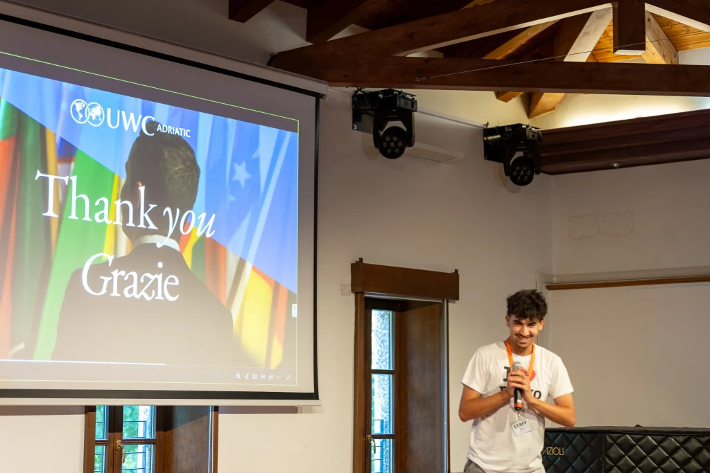 Un giovane in camicia bianca è in piedi accanto a uno schermo su cui è scritto "Thank you Grazie", con delle bandiere e il logo "UWC Adriatic". Sta sorridendo e tiene in mano un microfono in una stanza ben illuminata con travi a vista.