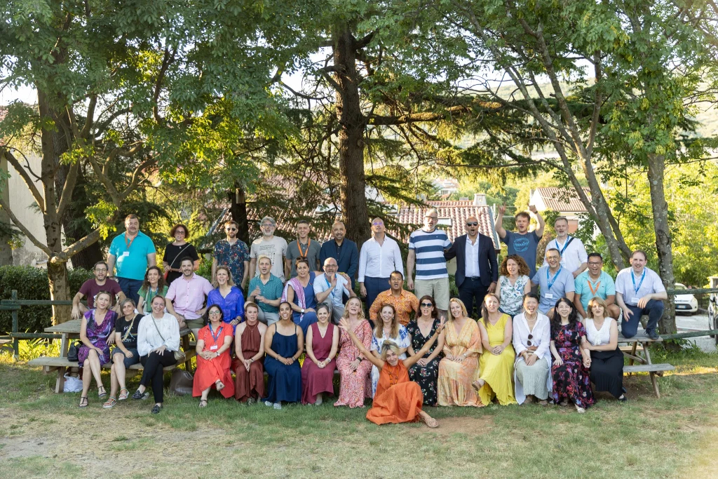 Un folto gruppo di persone posa all'aperto sotto gli alberi, seduto o in piedi su panchine e sull'erba, vestito con abiti colorati e semi-formali, sorridendo alla macchina fotografica con alcune case sullo sfondo.