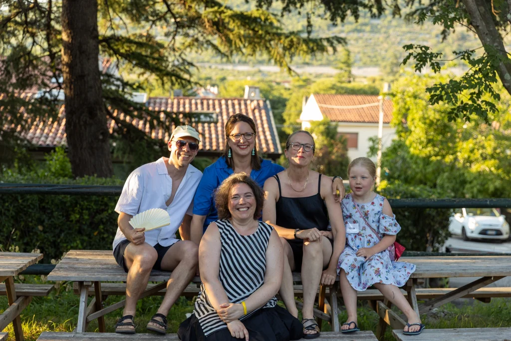 Cinque persone, tra cui un bambino, sono sedute su panchine di legno all’aperto, alla luce del sole, e sorridono alla macchina fotografica. Sullo sfondo si vedono alberi, case dai tetti rossi e vegetazione.