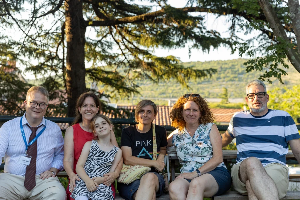 Sei adulti e un bambino sono seduti su una panchina all’aperto e sorridono alla macchina fotografica, con alberi e colline sullo sfondo in una giornata soleggiata.