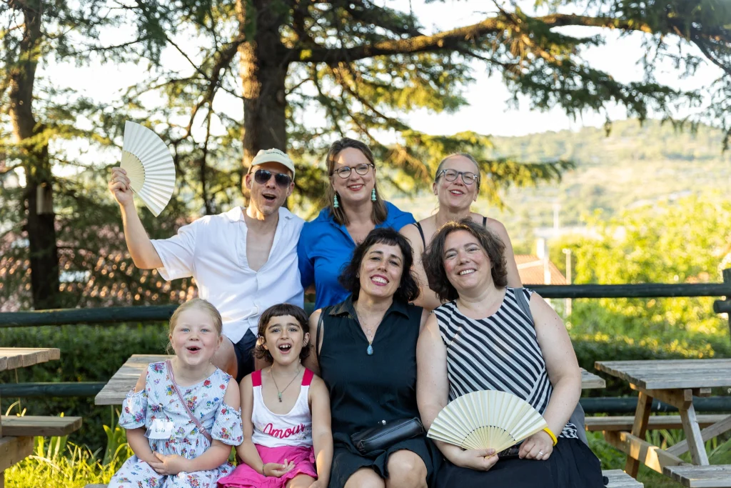 Un gruppo di sette persone, composto da quattro adulti e tre bambini, è seduto all’aperto su panchine di legno e sorride, con in mano dei ventagli, mentre sullo sfondo si vedono alberi e la luce del sole.