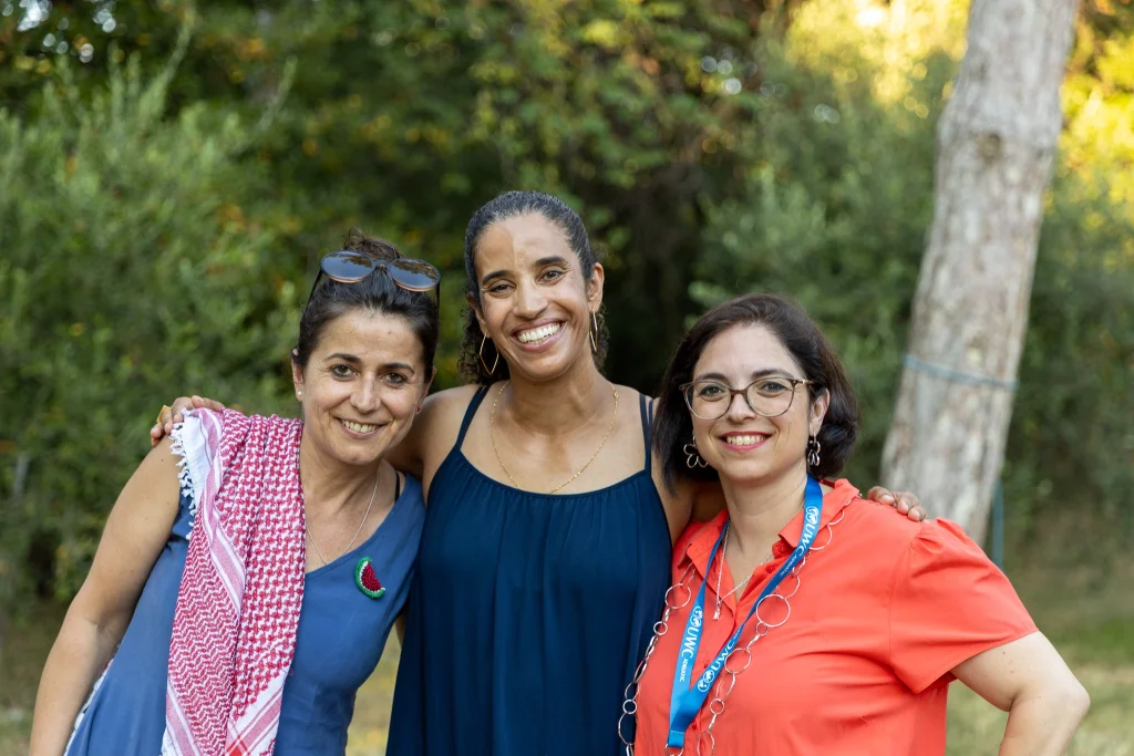Tre donne sono in piedi all'aperto e sorridono alla fotocamera. La donna a sinistra indossa occhiali da sole e una sciarpa a fantasia, quella al centro indossa un abito blu scuro, mentre quella a destra indossa occhiali da vista e un top color corallo. Sullo sfondo si vedono degli alberi.