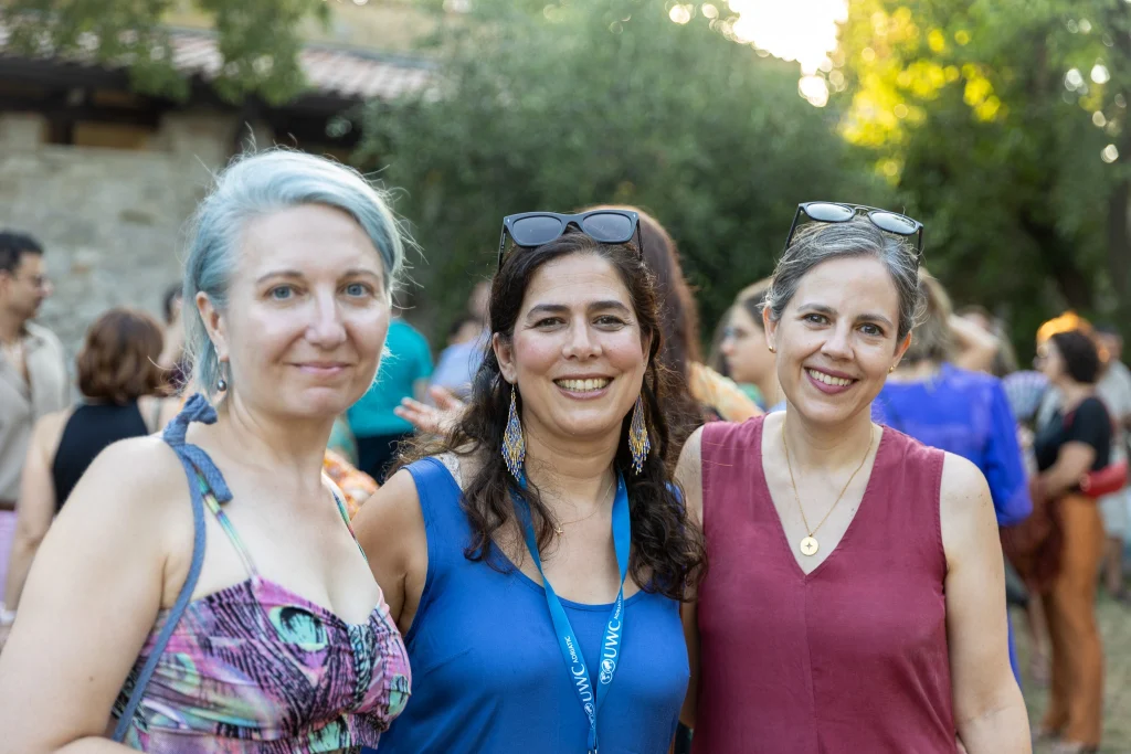 Tre donne sorridono alla fotocamera durante un raduno all'aperto, in piedi una accanto all'altra. Sullo sfondo sfocato si intravedono persone e alberi, a suggerire un evento informale e soleggiato.