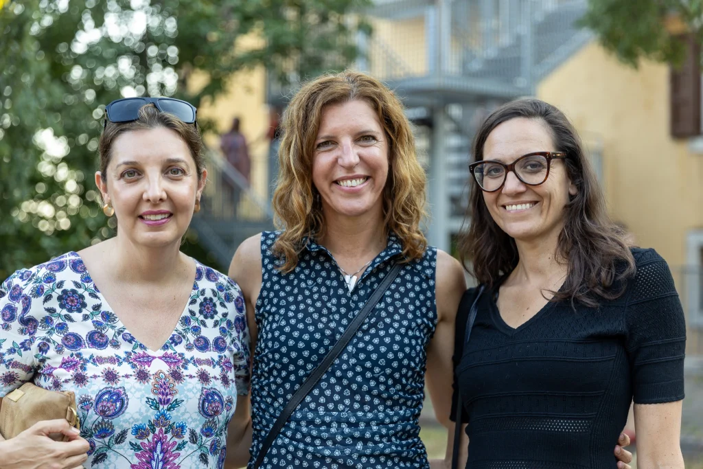 Tre donne sono in piedi all'aperto, vicine l'una all'altra, e sorridono alla fotocamera. Sullo sfondo si vedono degli alberi e un edificio. Le donne indossano abiti casual e sembrano godersi una piacevole giornata.