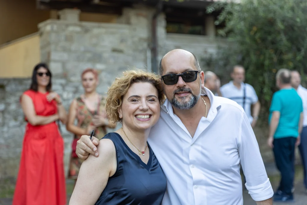 Una donna e un uomo sorridenti, entrambi con gli occhiali da sole, sono in piedi vicini all'aperto. Sullo sfondo si intravedono diverse persone sfocate, alcune vestite di rosso e altre in abiti casual, vicino a un edificio in pietra.