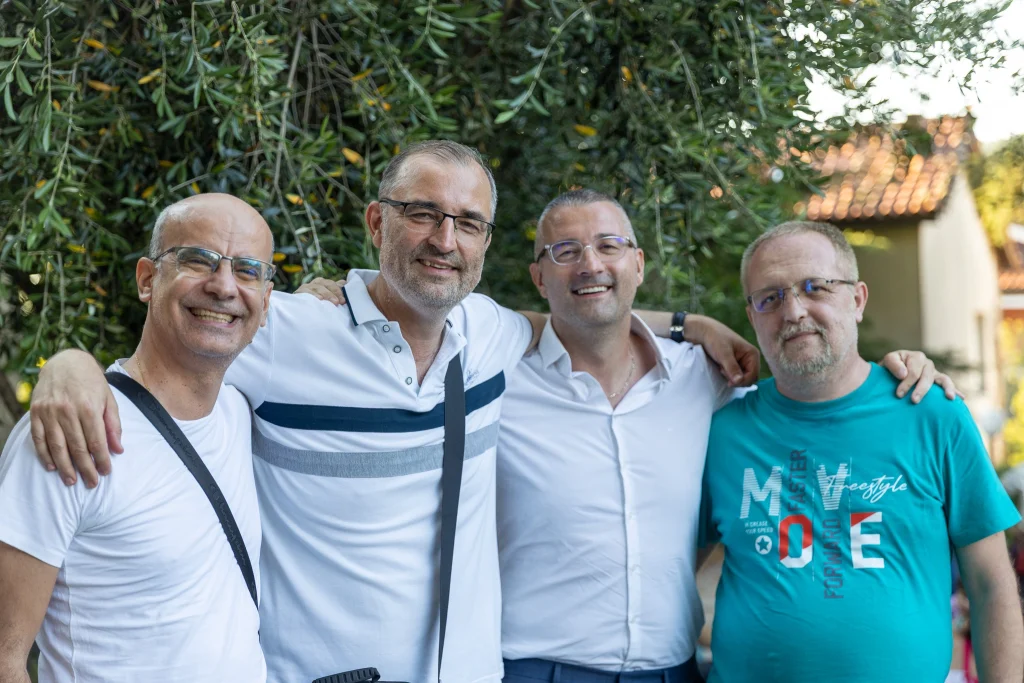 Quattro uomini sorridenti sono in piedi all’aperto, con le braccia intrecciate, mentre posano per una foto di gruppo vicino ad alberi rigogliosi, con una casa dal tetto di tegole sullo sfondo.