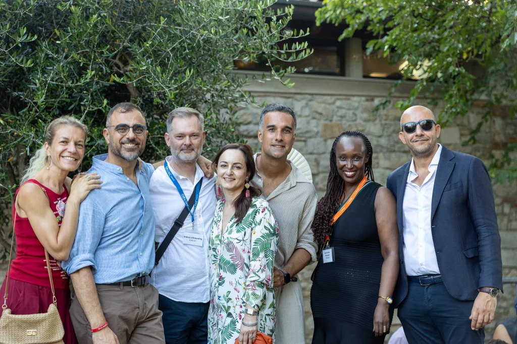 Sette persone sono in piedi all'aperto, vicine l'una all'altra, e sorridono alla fotocamera. Indossano abiti estivi, con del verde e un edificio in pietra sullo sfondo. Il gruppo sembra rilassato e felice.