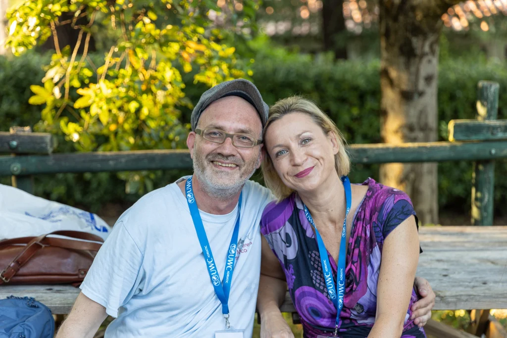 Un uomo e una donna sorridenti sono seduti vicini su una panchina all'aperto; entrambi indossano badge identificativi e cordoncini blu. Sullo sfondo si vedono alberi e la luce del sole.