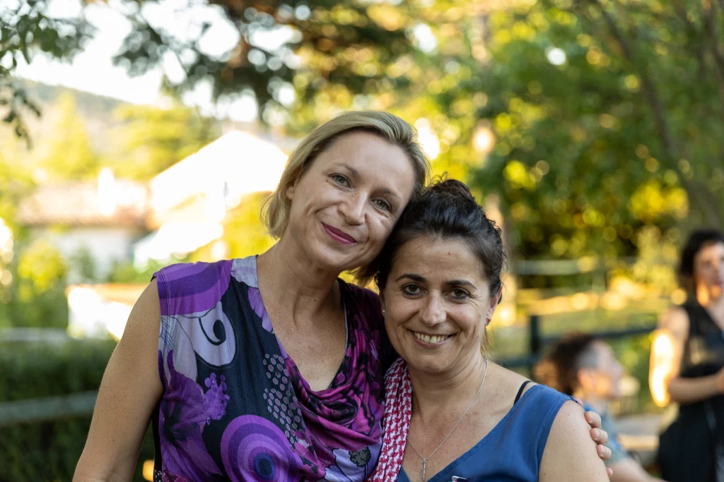 Due donne in piedi una accanto all'altra all'aperto, che sorridono alla fotocamera. Lo sfondo è ricco di vegetazione e luce solare, con altre persone sfocate in lontananza.