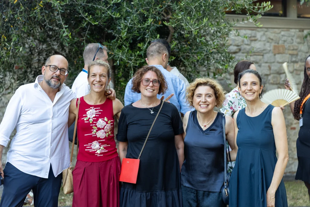 Cinque persone sono in piedi all'aperto e sorridono per una foto di gruppo davanti a una vegetazione rigogliosa e a un muro di pietra. In primo piano ci sono quattro donne e un uomo; sullo sfondo si intravedono altre persone.