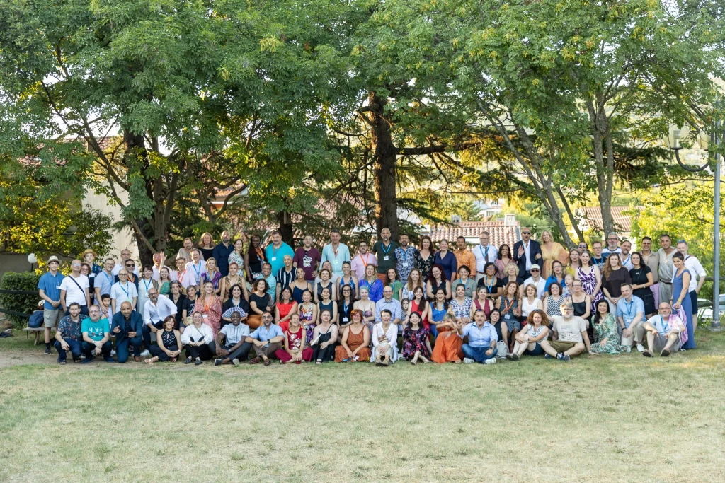 Un folto gruppo di persone posa insieme all'aperto, sul prato davanti ad alcuni alberi; tutti guardano verso la fotocamera e sorridono. Il gruppo è composto da adulti e bambini vestiti in modo informale e semi-formale.