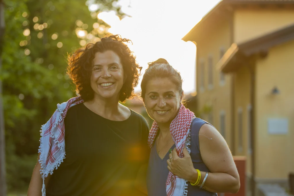 Due donne sorridenti all'aperto al tramonto, in piedi una accanto all'altra con scialli a motivi geometrici drappeggiati sulle spalle. Sullo sfondo si vedono un edificio giallo e alberi verdi, con la luce del sole che crea un effetto caldo e luminoso.