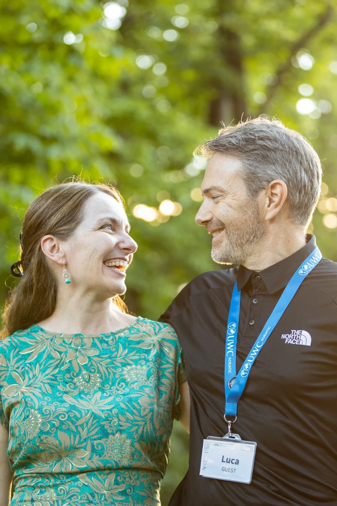 Una donna con un vestito verde a fantasia e un uomo con un cordino blu si scambiano un sorriso all'aperto, circondati dal verde e dalla luce del sole.