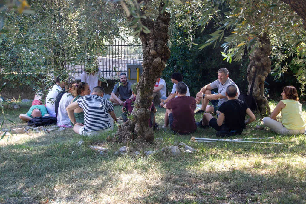 Un gruppo di persone è seduto in cerchio sull'erba sotto gli alberi, intento a conversare. La luce del sole filtra tra le foglie e sullo sfondo si intravede una recinzione. Alcune persone sono sedute per terra, altre sono appoggiate ai tronchi degli alberi.