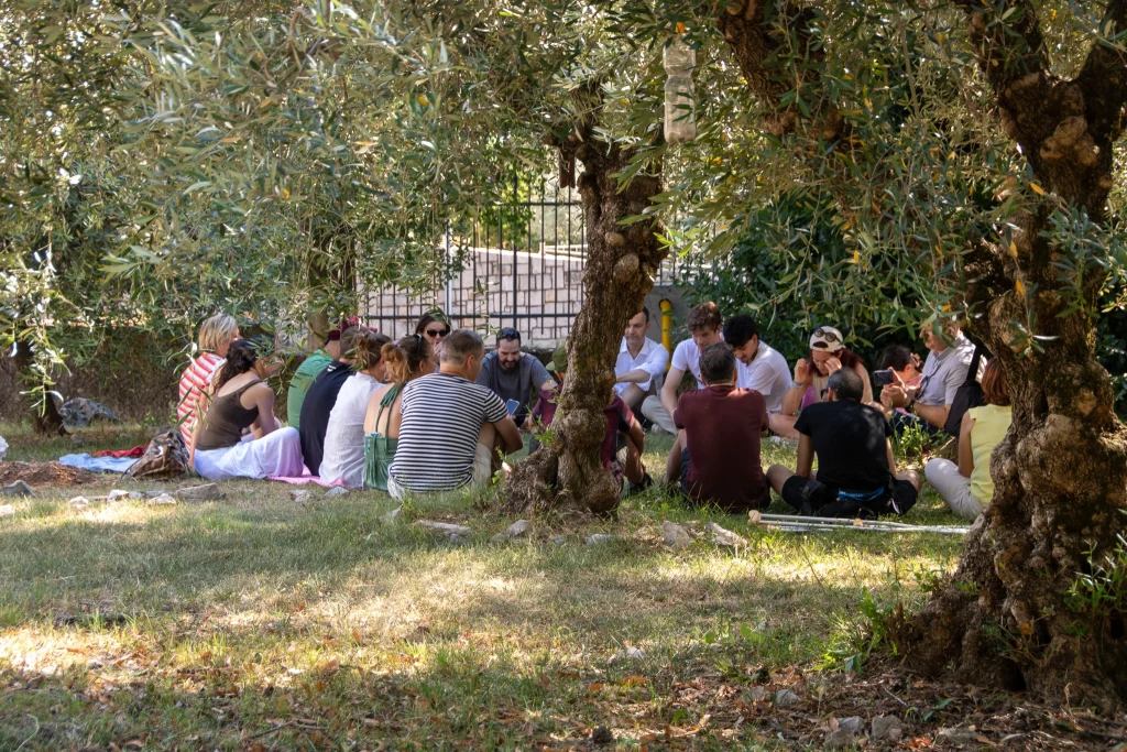 Un gruppo di persone è seduto in cerchio sull'erba all'ombra degli alberi, intento a conversare o a discutere all'aperto in una giornata di sole.